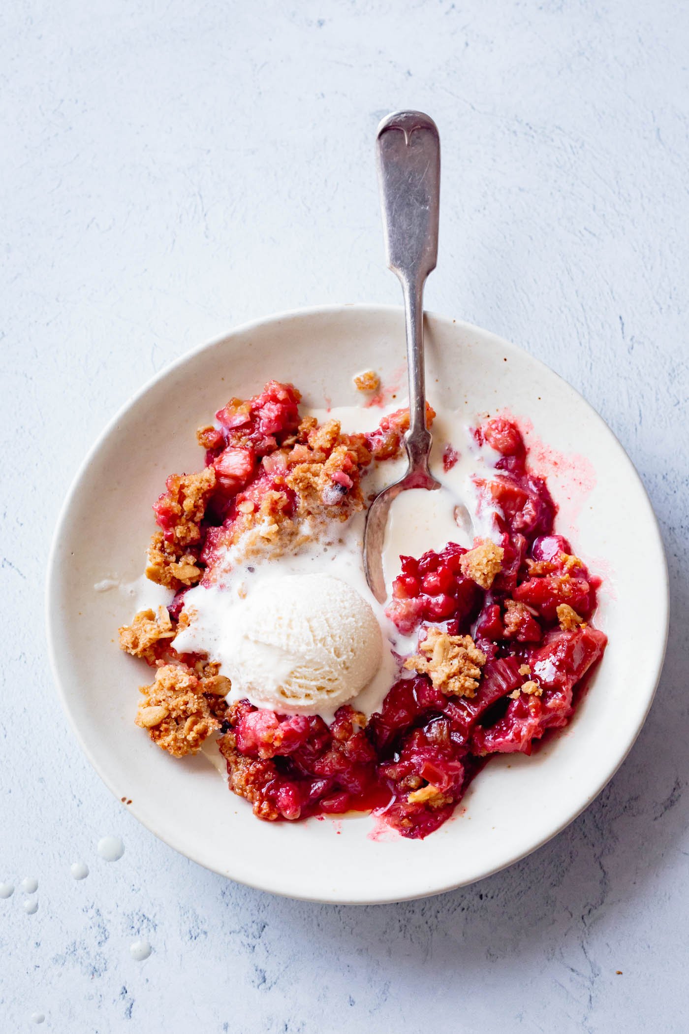 a spoon sits in a bowl of strawberry rhubarb crisp topped with gooey melty ice cream
