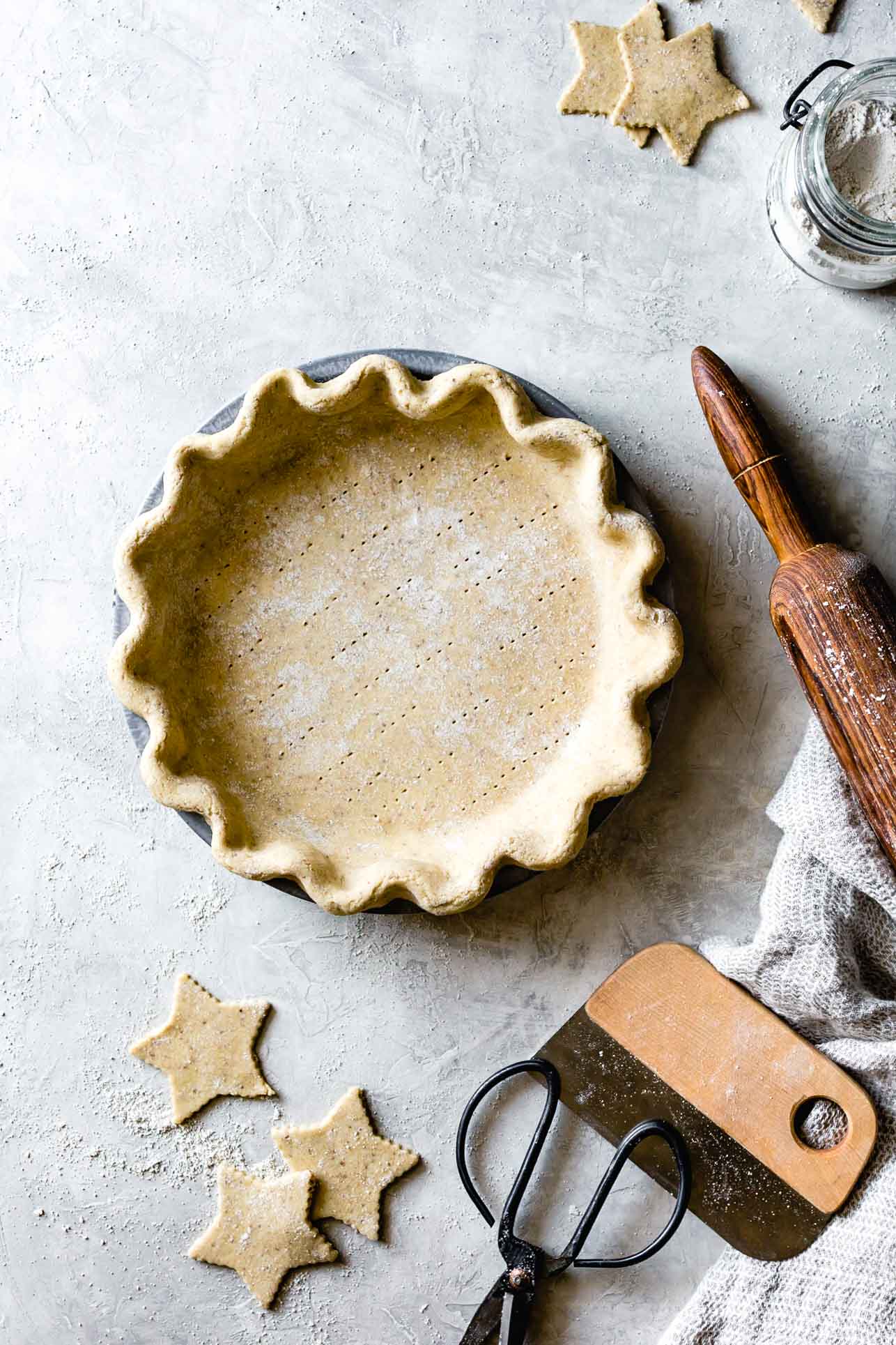 A handsome, unbaked gluten-free pie crust sits on a plaster surface with star cut-outs, a rolling pin, and a gray dish towel around it