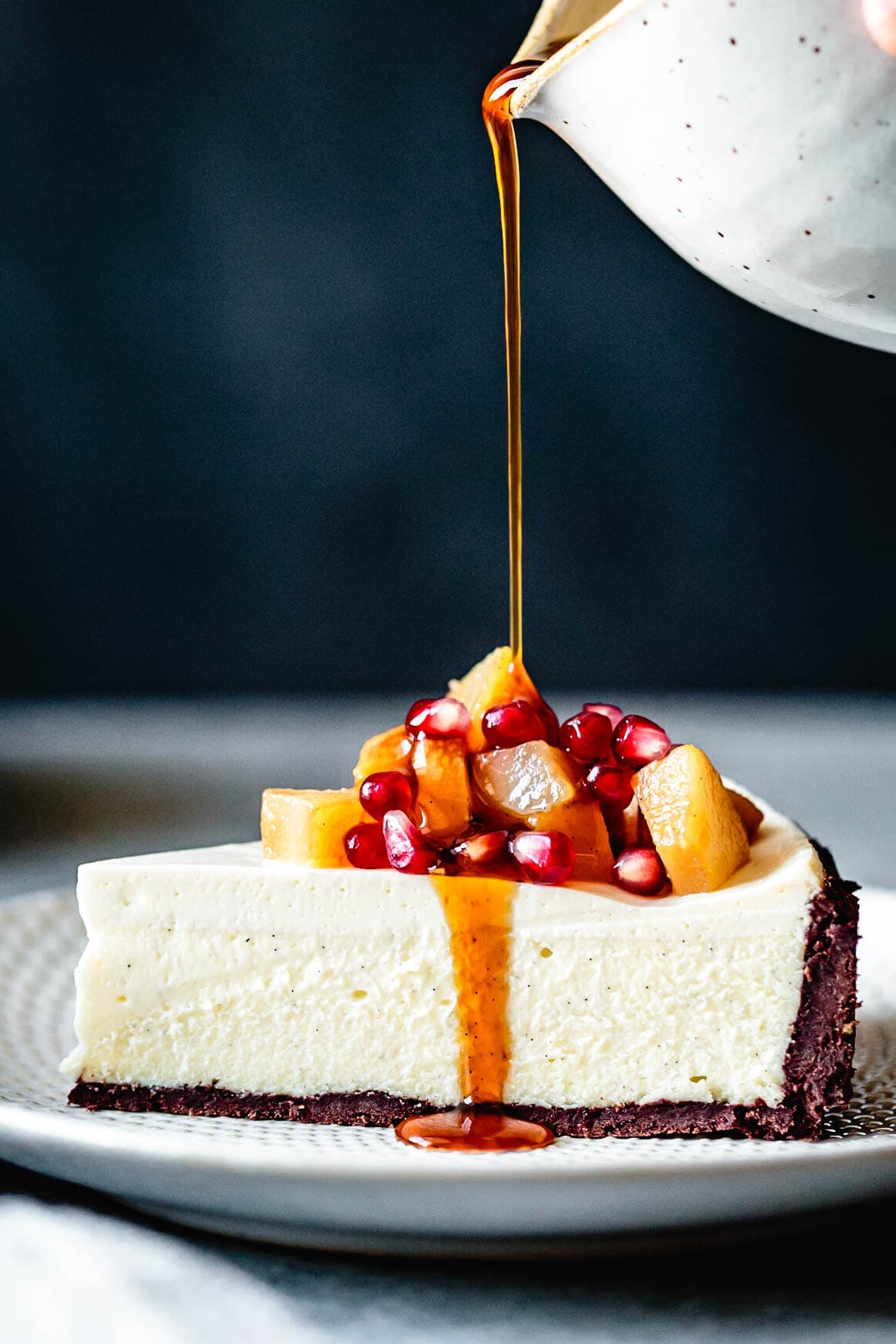 a slice of goat cheese cheesecake shown from the side is being smothered in golden syrup, poached pears, and pomegranate seeds