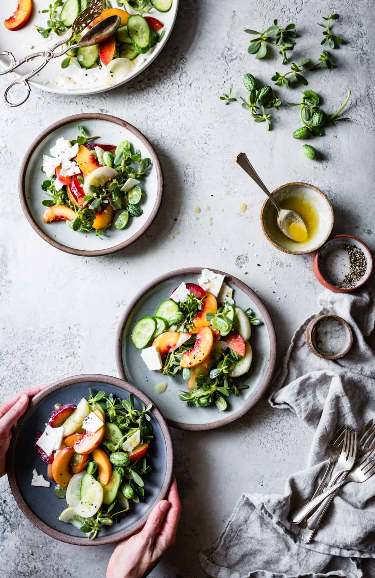 hands holding a plate of Stone Fruit Cucumber Salad with Ricotta Salata, Purslane, and Golden Balsamic Miso Vinaigrette