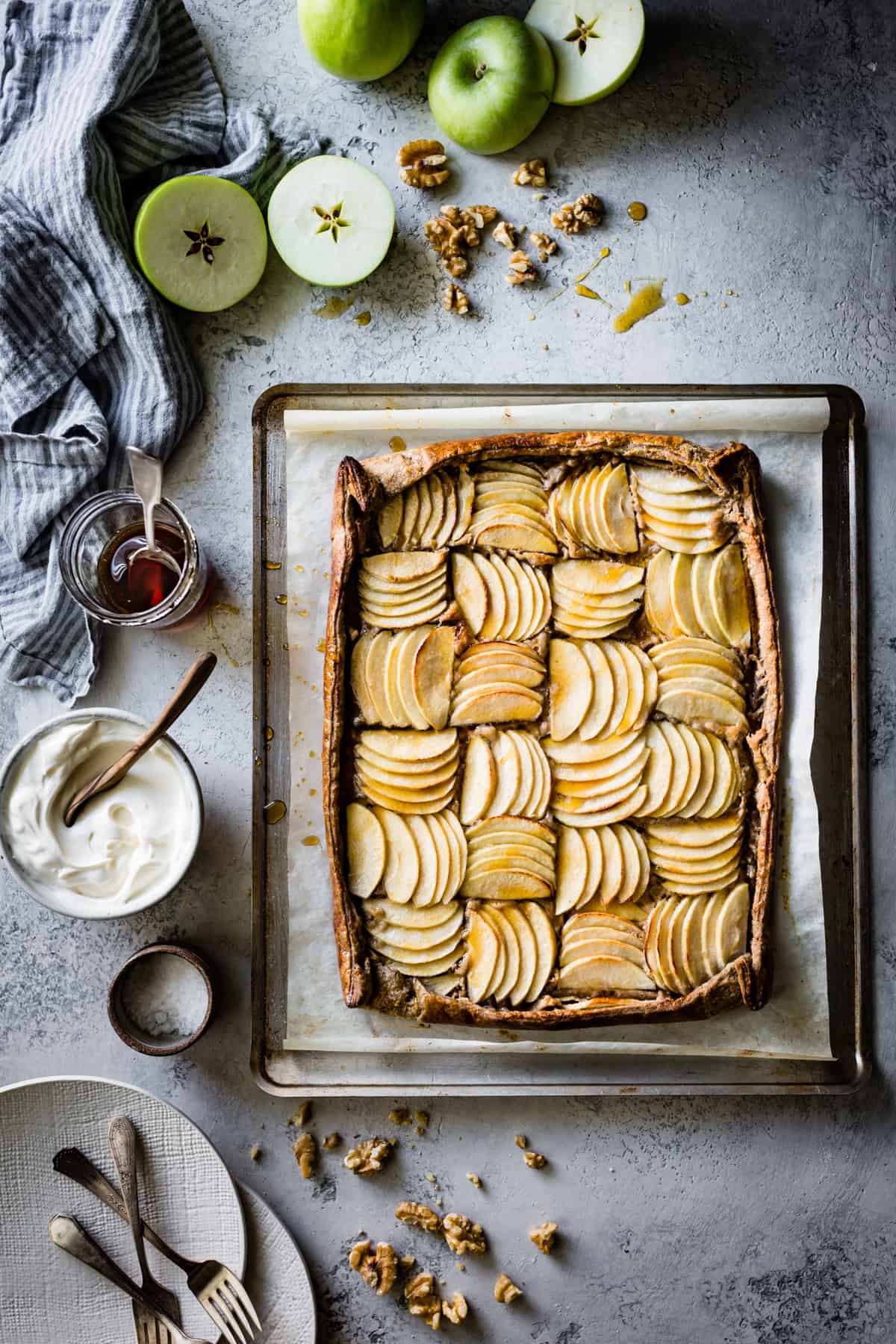 Buckwheat Apple Galette on a gray surface with apples, cream, and walnuts scattered around