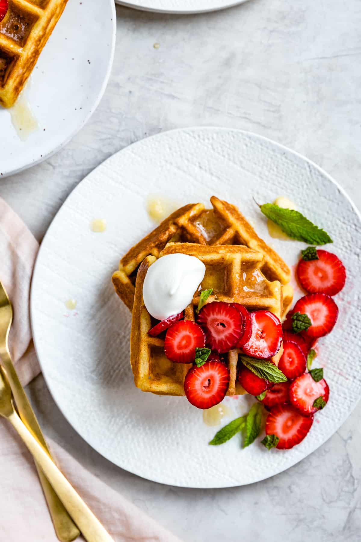 two corn flour waffles are stacked on a textured white plate and topped with glossy strawberries and mint
