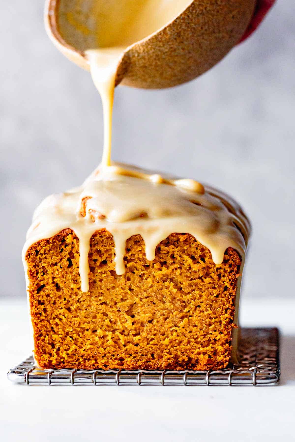 maple glaze is being drizzled over a golden loaf of pumpkin bread with a floofy crumb