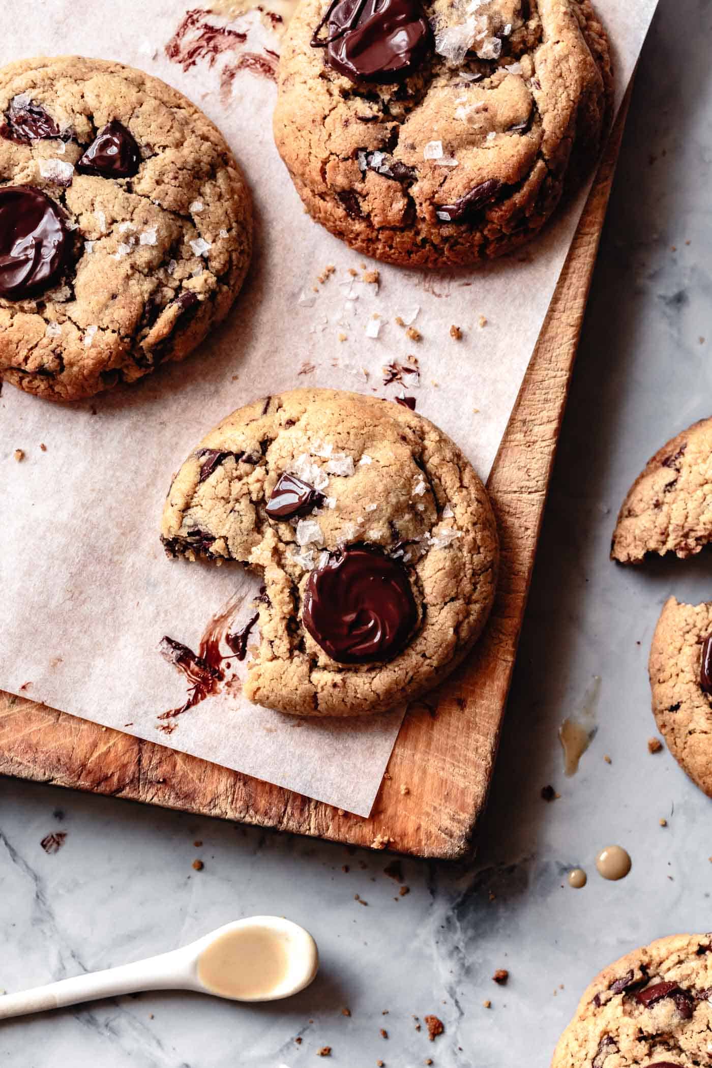 A tahini chocolate chunk cookie with a bite taken out sits on a wooden board