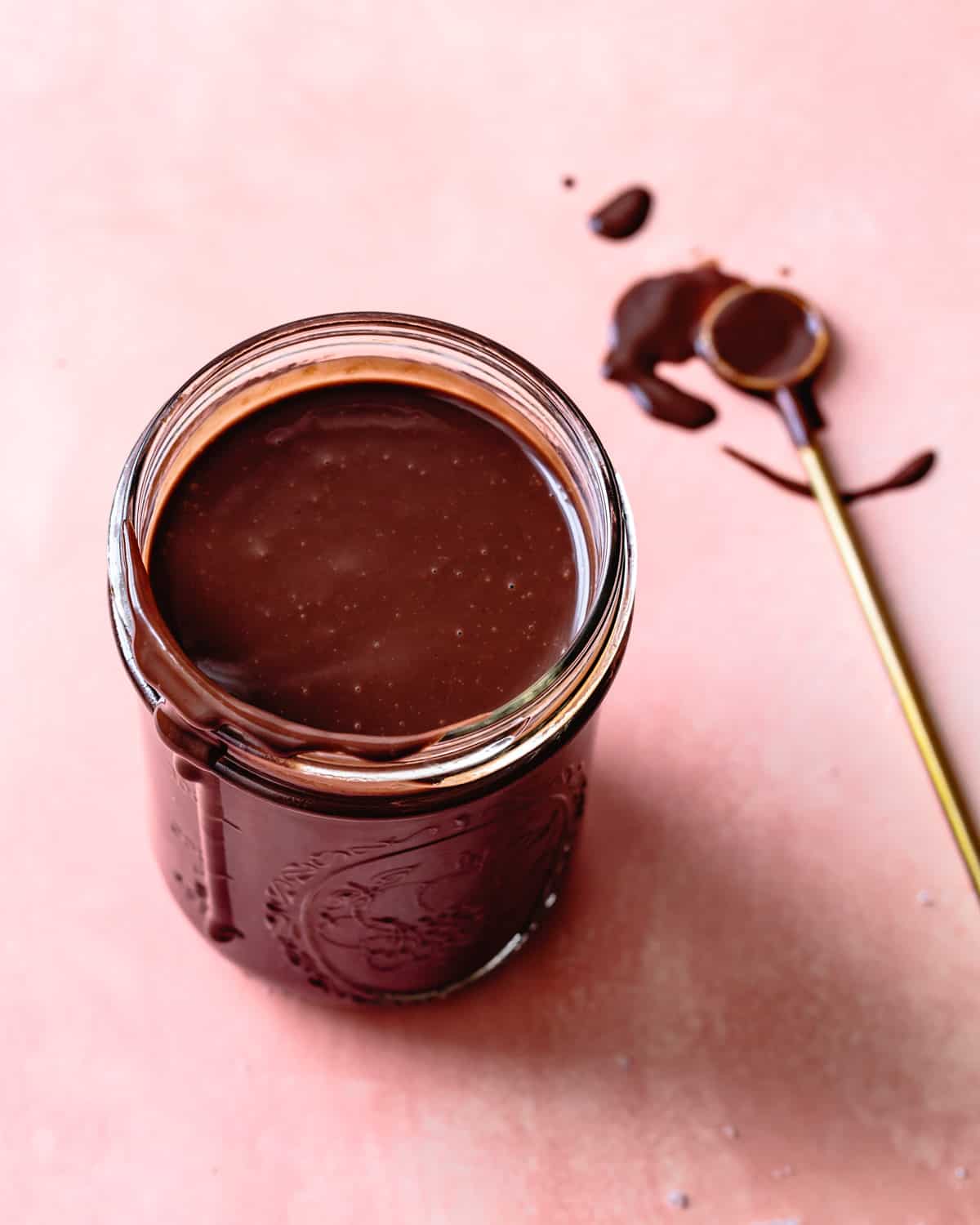 jar of hot fudge sauce on pink background with gold spoon