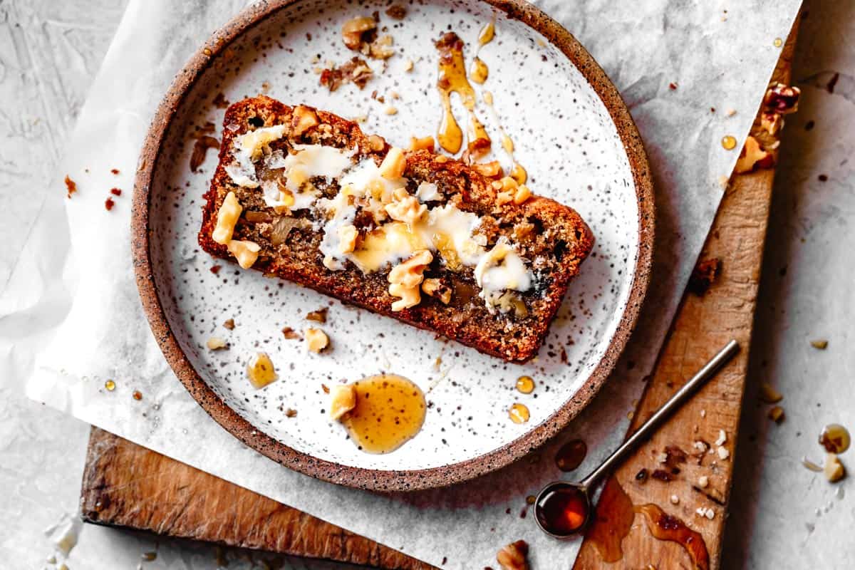 slice of healthy banana bread on a plate with butter, honey, and walnuts