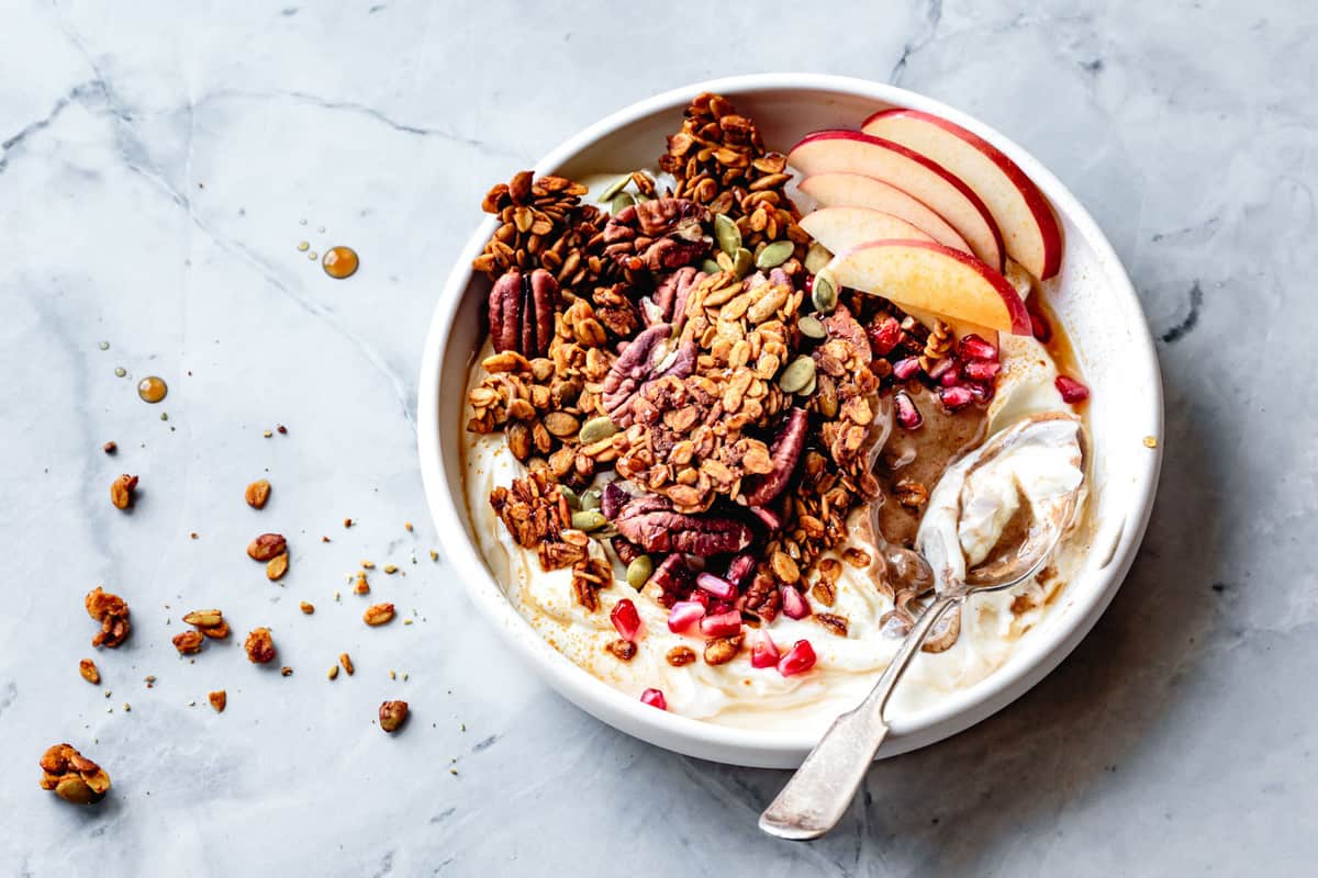pumpkin pie granola in a bowl with yogurt, nut butter, apple slices, and pomegranate arils