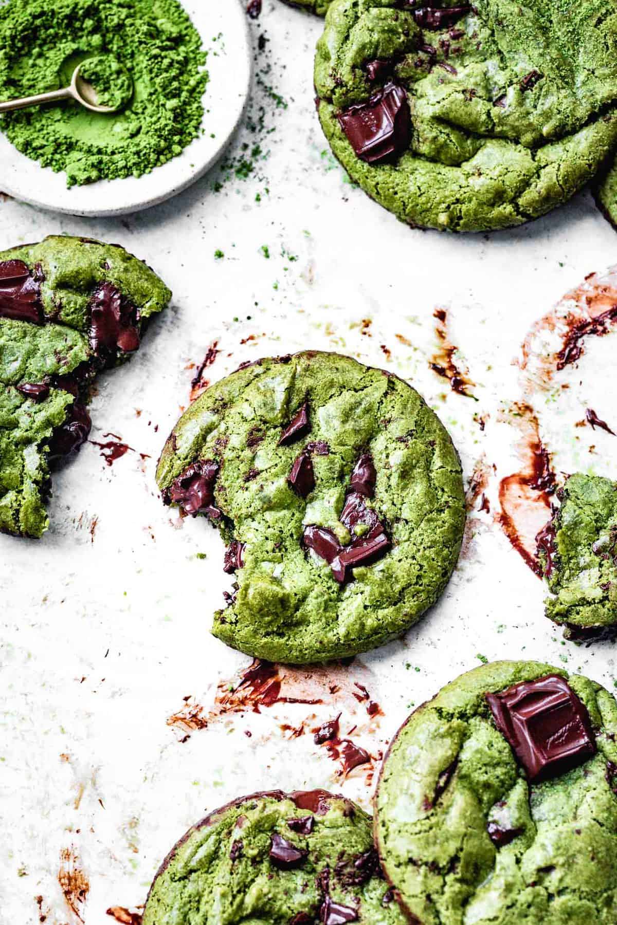 a messy piece of parchment smeared with chocolate has several gooey matcha chocolate chip cookies on it, one with a tantalizing bite taken out
