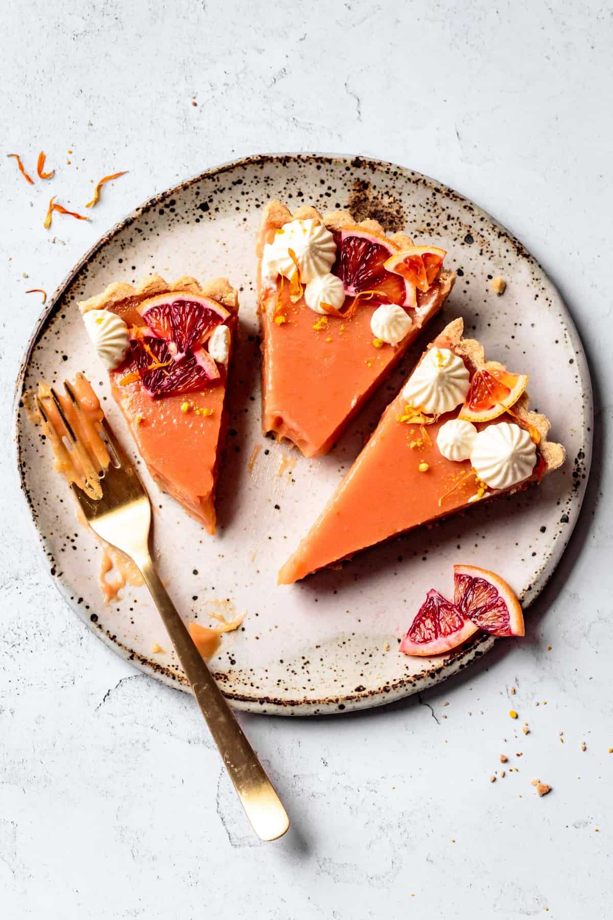 vertical overhead shot of a speckled plate with three blood orange tart slices on it and a gold fork