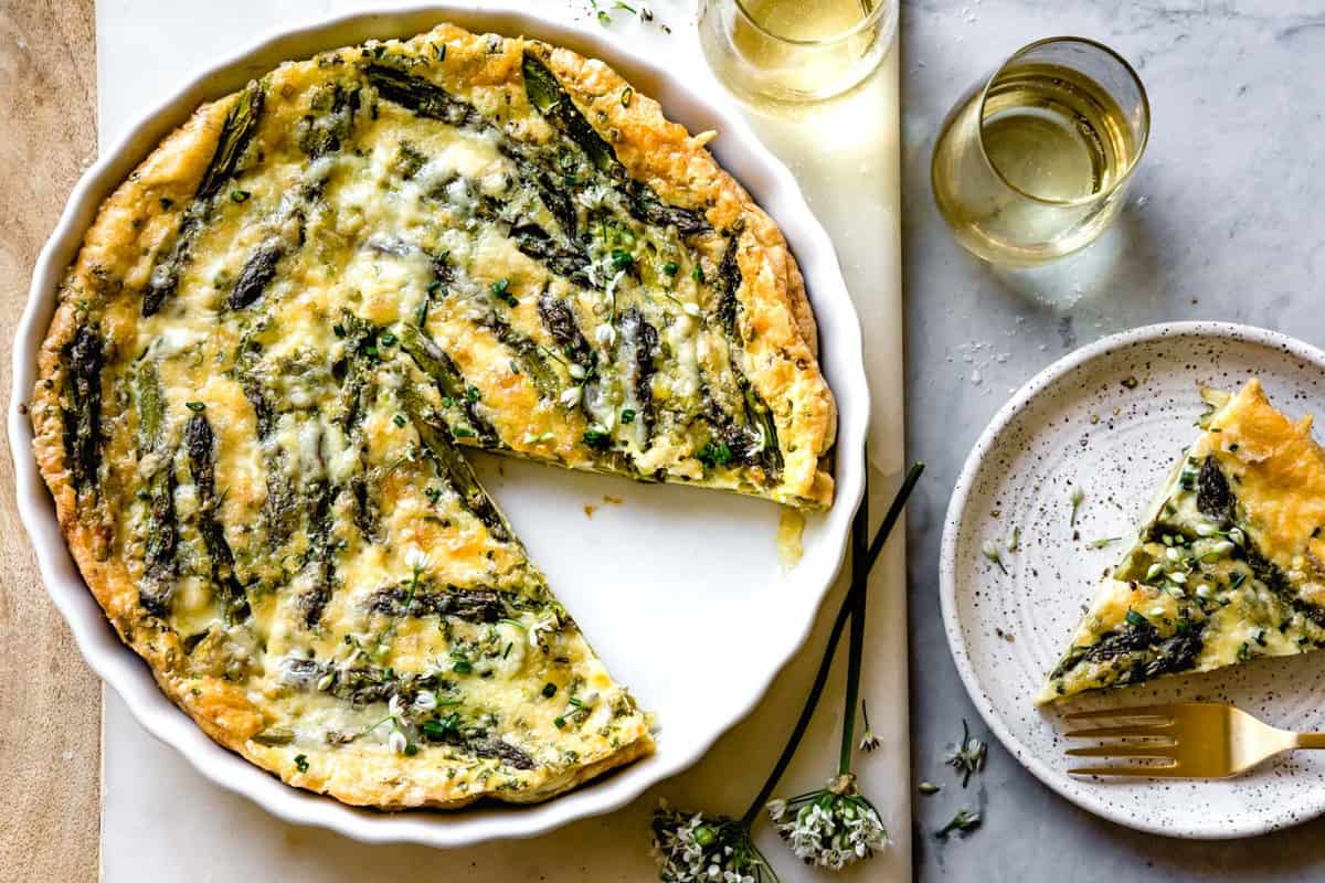 baked quiche on a marble board with a slice cut out and glasses of white wine