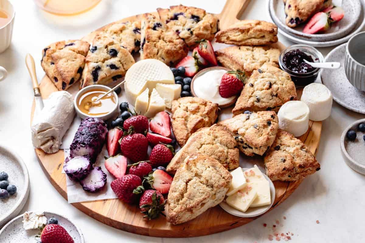 a wide shot of a beautiful spread of cheeses, fruit, scones, butter, and honey on a round wooden board