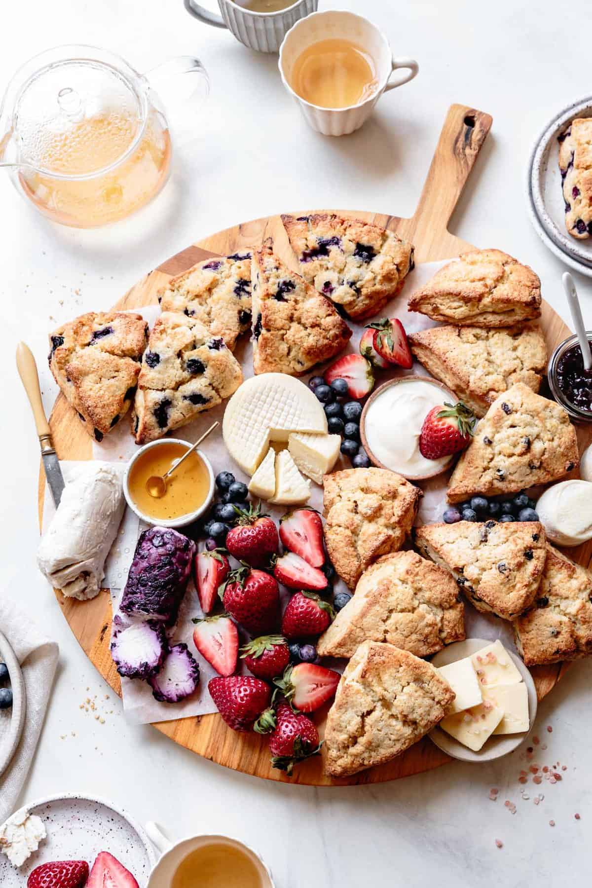 a beautiful spread of cheeses, fruit, scones, butter, and honey on a round wooden board