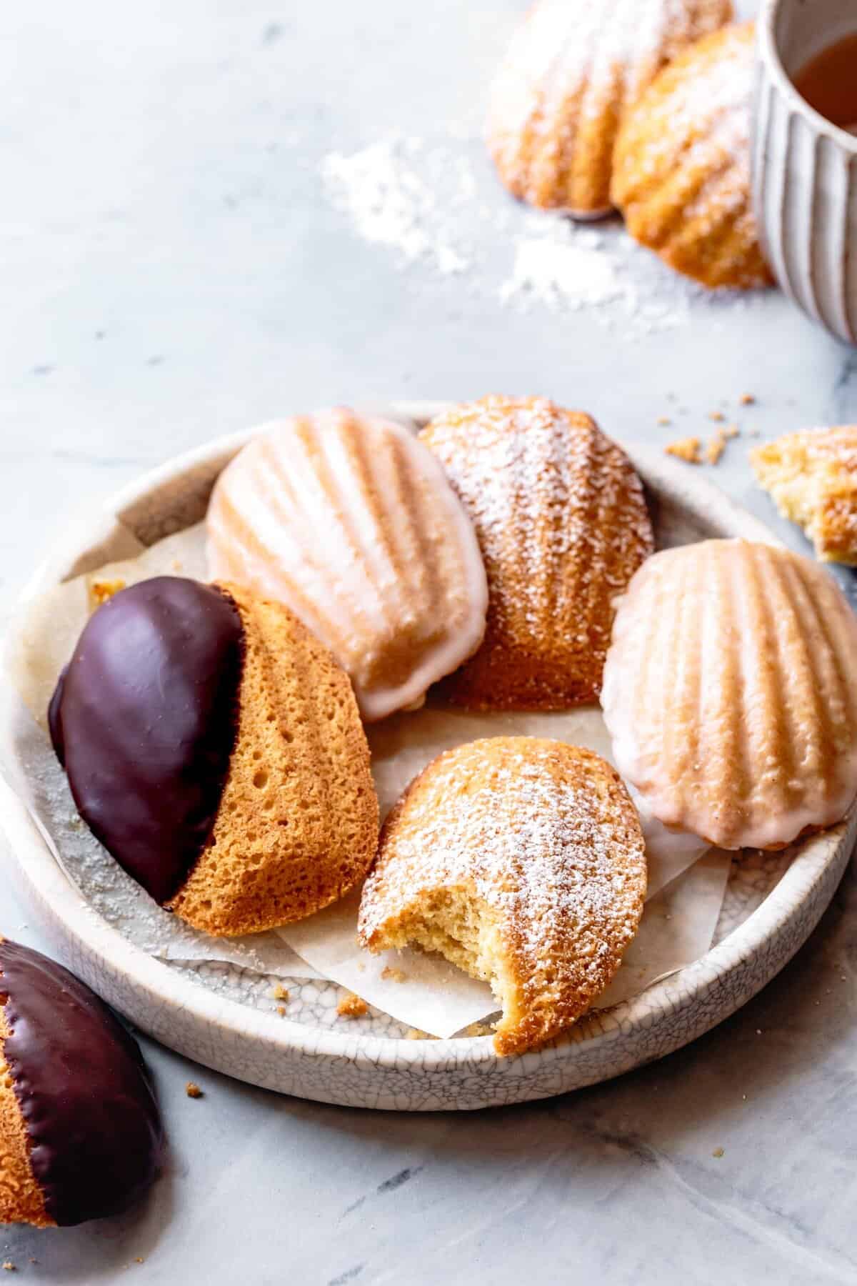 five GF madeleines on a plate with different toppings (chocolate, lemon glaze, and powdered sugar), one with a bite taken out to show the spongey interior