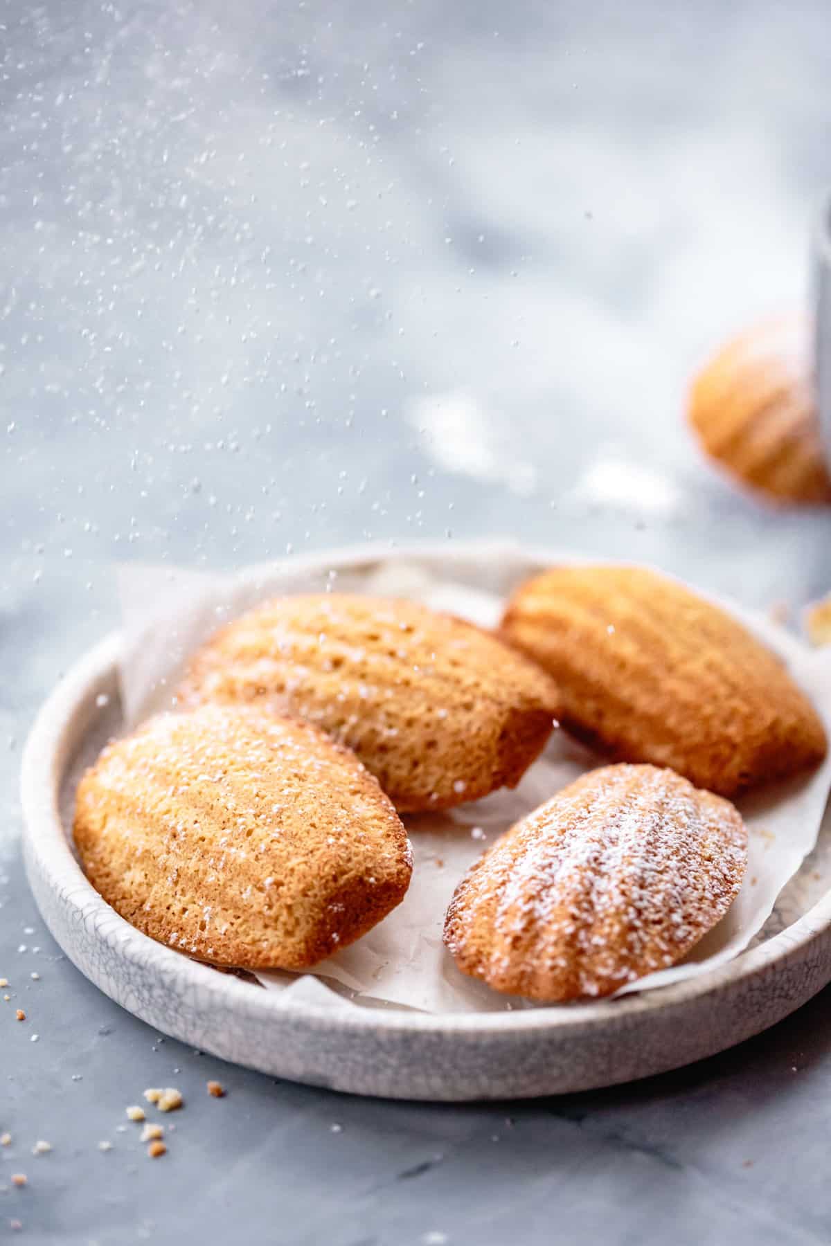 madeleines on a plate being sprinkled with powdered sugar in a dreamy bokeh