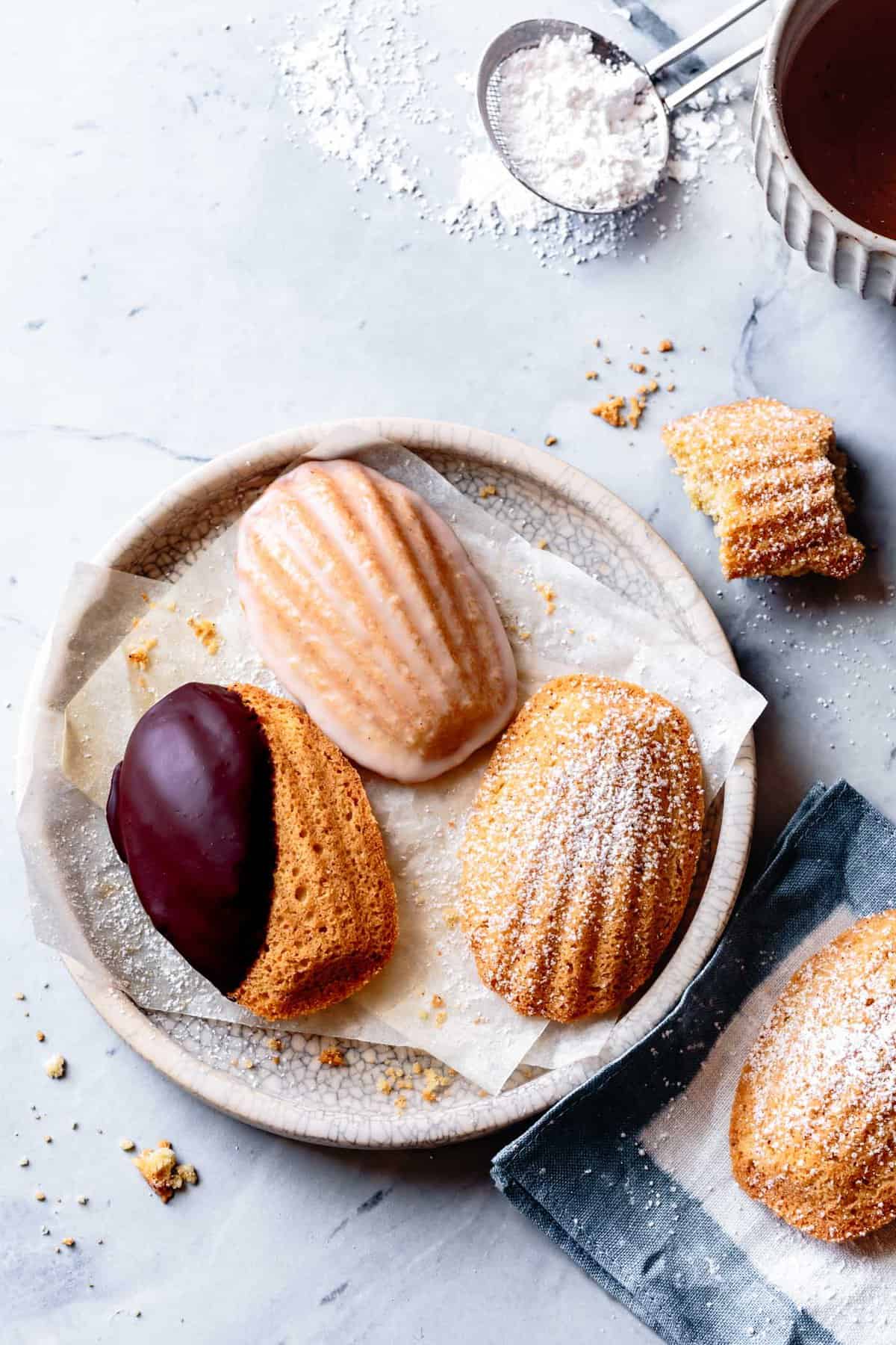 three gluten-free madeleines on a plate with crumbs and a napkin on a marble surface