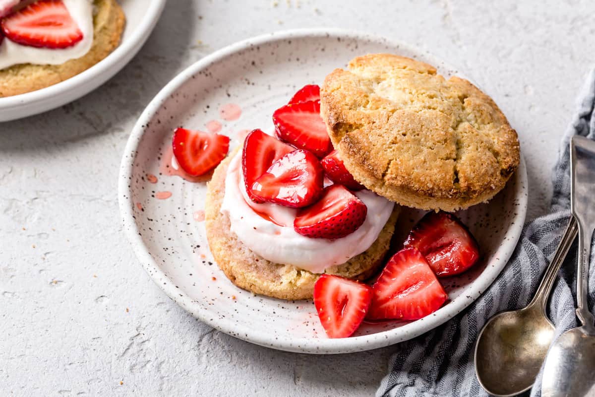 Wide, three-quarter angle shot of a shortcake on a cute speckled white plate with two spoons and a linen next to it. 