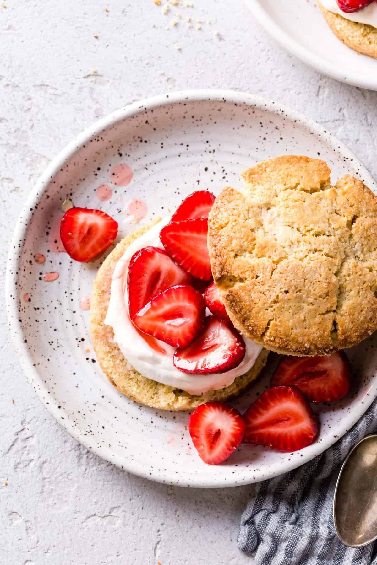 A gluten-free strawberry shortcake on a plate with glistening strawberries, swirls of cream, and a craggy golden biscuit