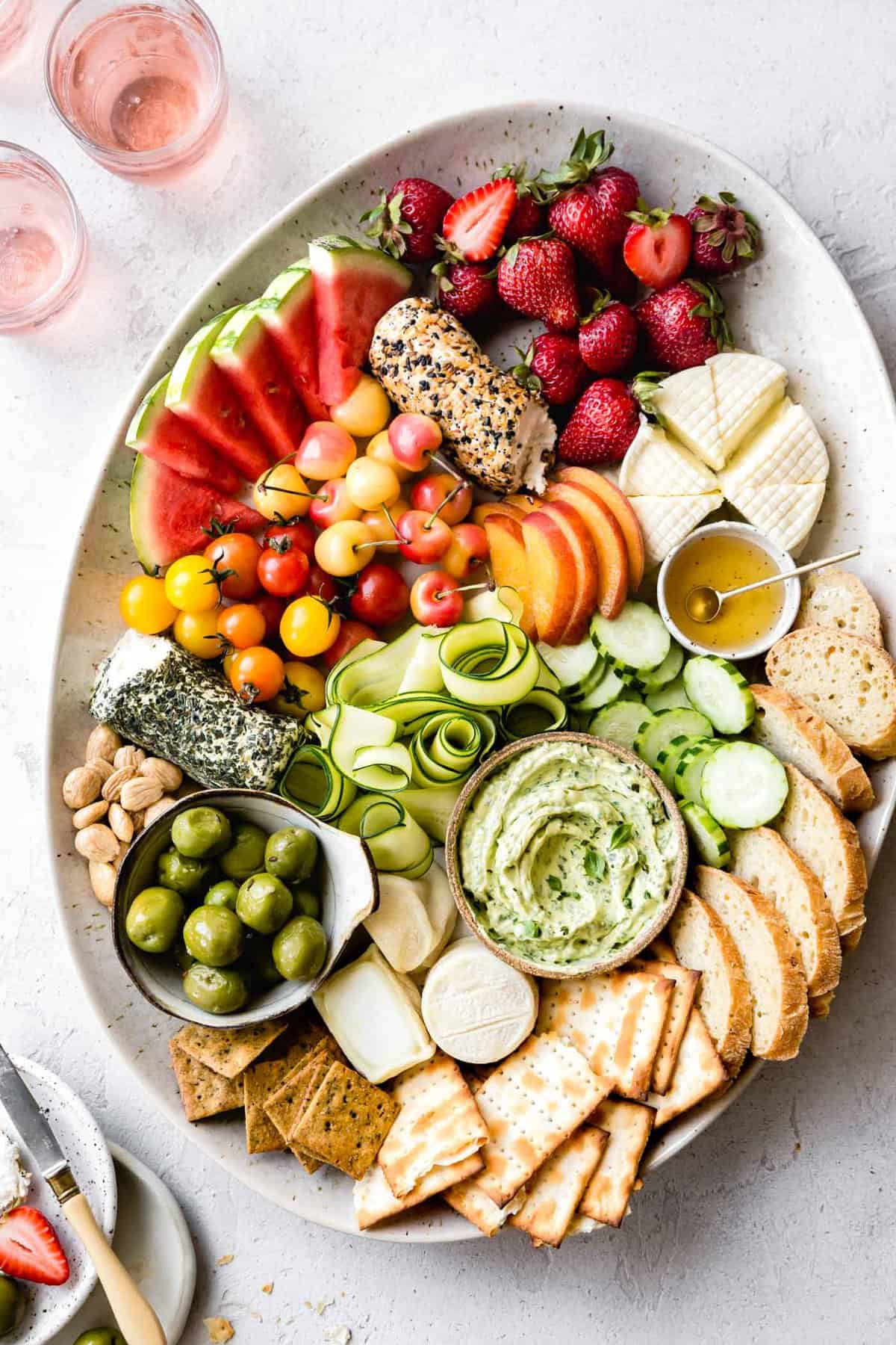 An oval platter is loaded with a rainbow of vibrant summer fruits and vegetables, cheeses, bread, and crackers. 