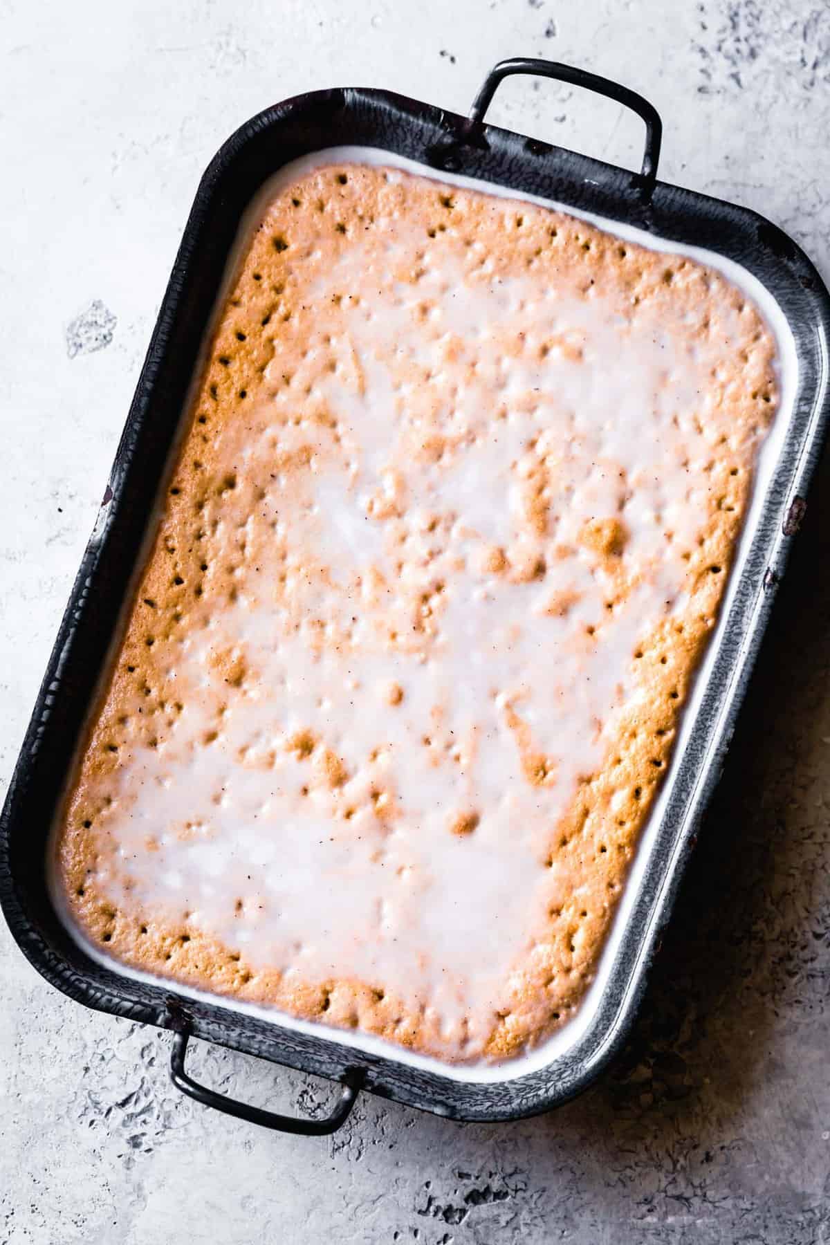 Tres leches cake in the pan with holes poked in the cake and creamy vanilla-flecked milk mixture pooling on top