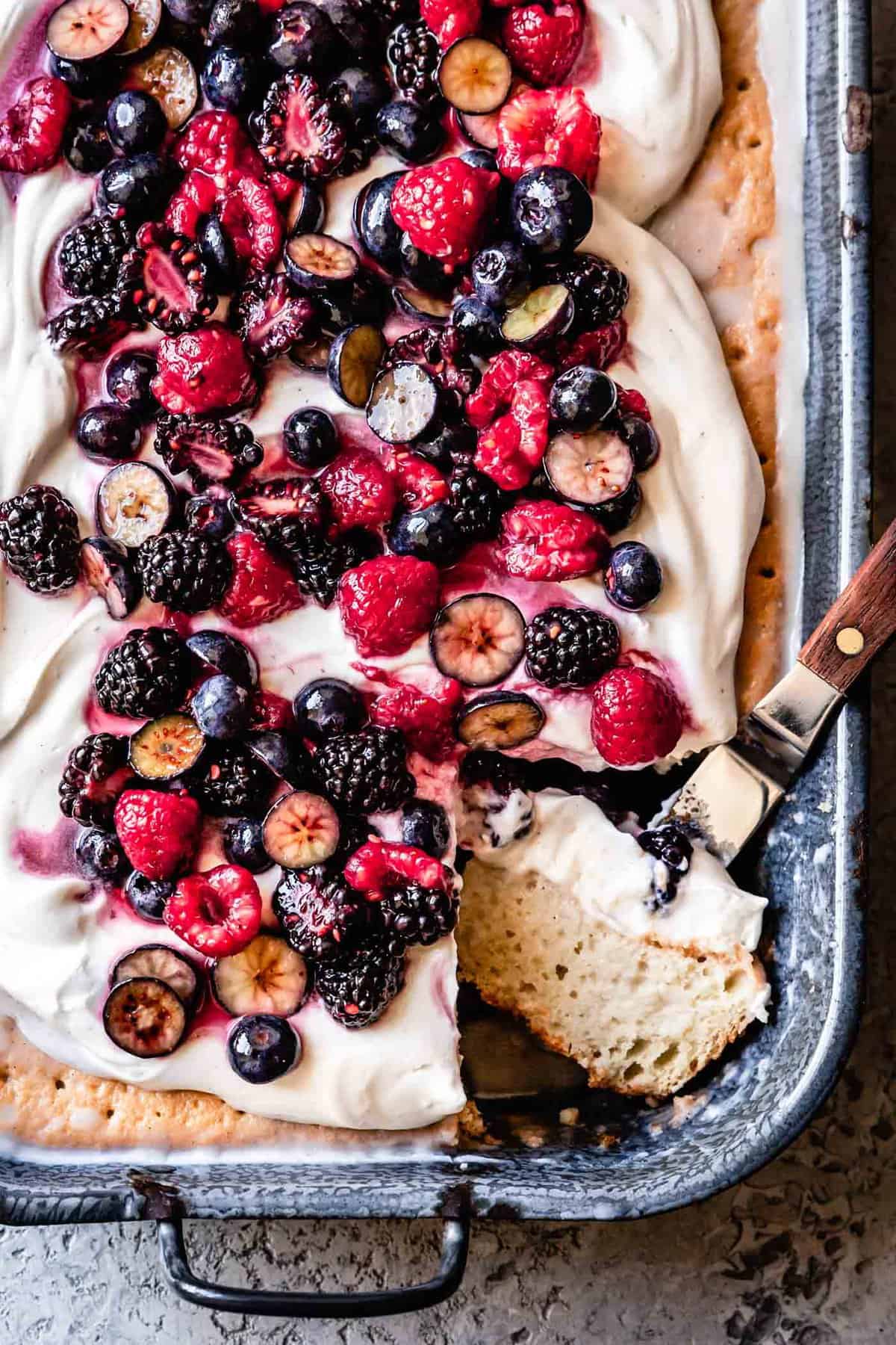 a gluten-free tres leches cake sits in a gray-blue graniteware pan topped with floofy whipped cream and juicy berries