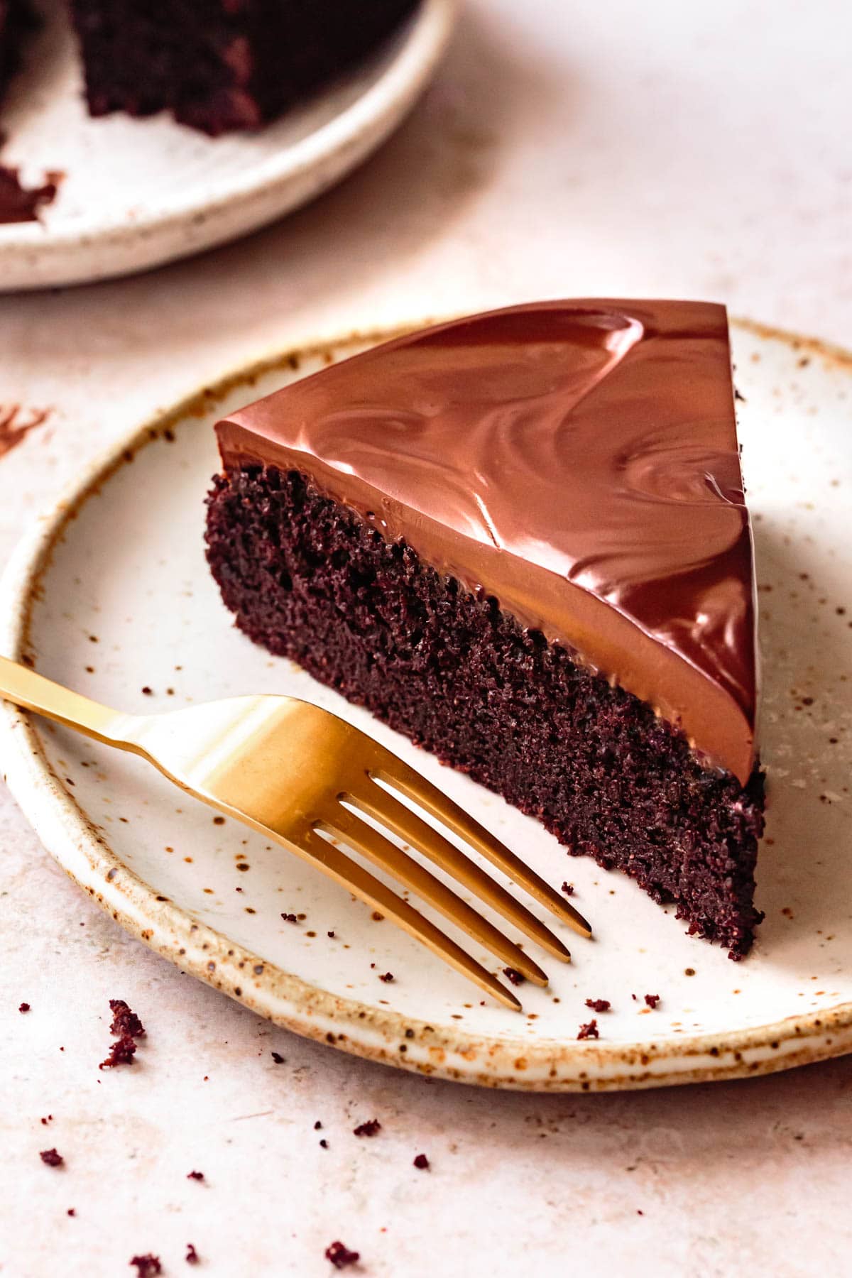 a handsome slice of chocolate cake crowned in coconut milk chocolate ganache sits on a speckled beige plate with a gold fork ready to dig in