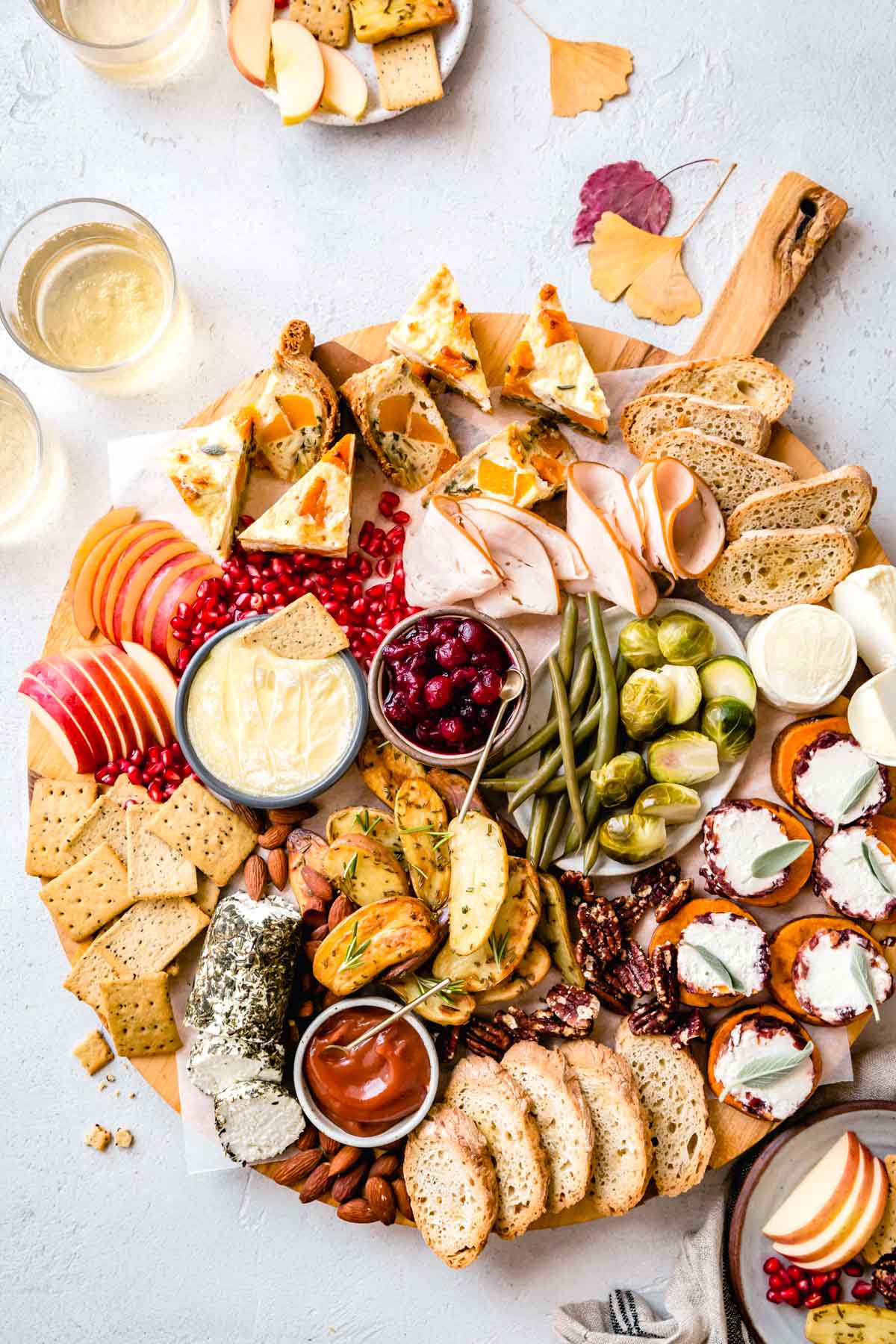 A fall harvest cheese board replete with cheeses, quiche pieces, bread, turkey, pickled veggies, cranberries, apples, sweet potato bites, and roasted potatoes