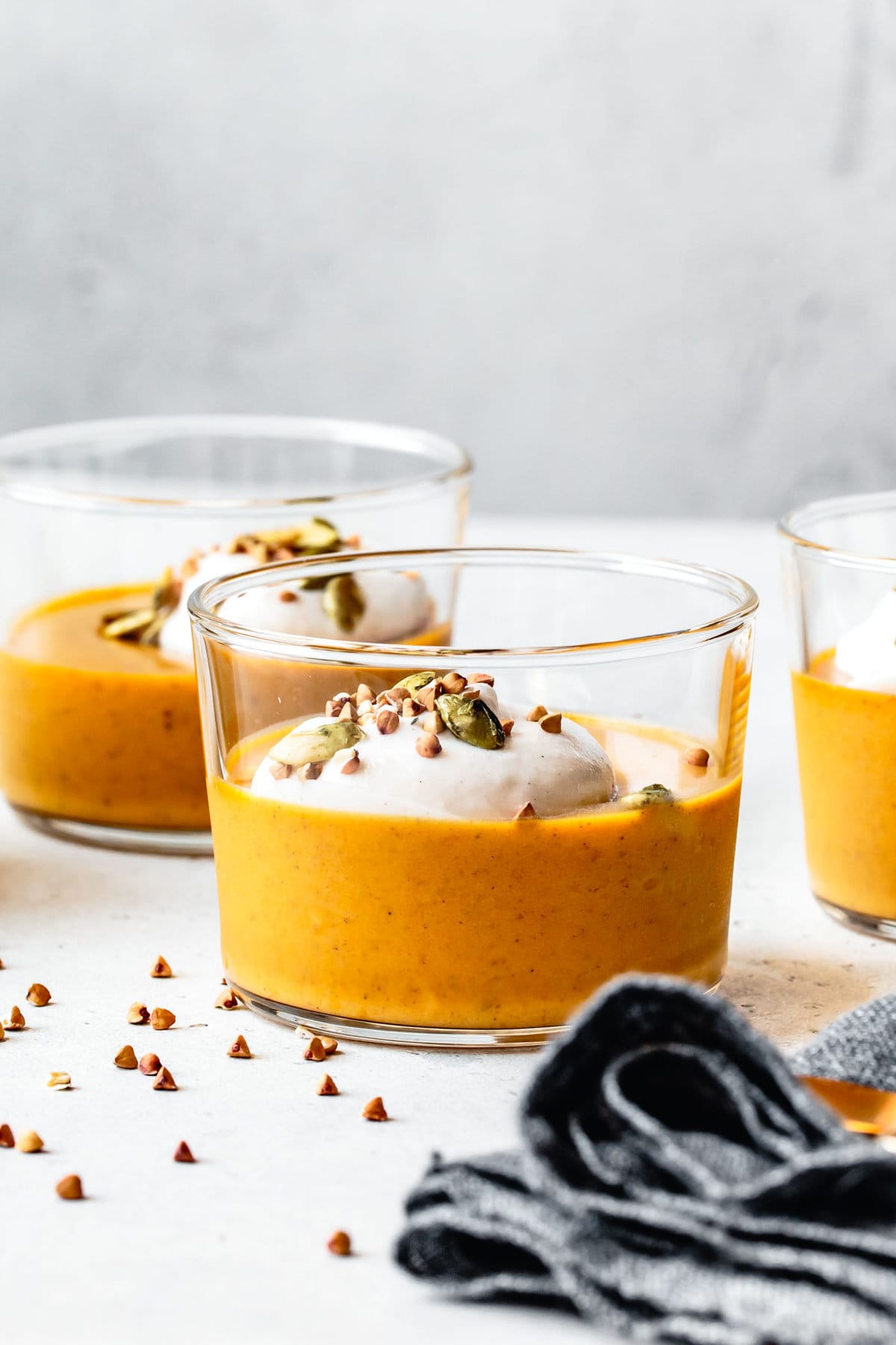 jars of golden pumpkin pudding sit on a light colored surface topped with plumes of cream and buckwheat groats
