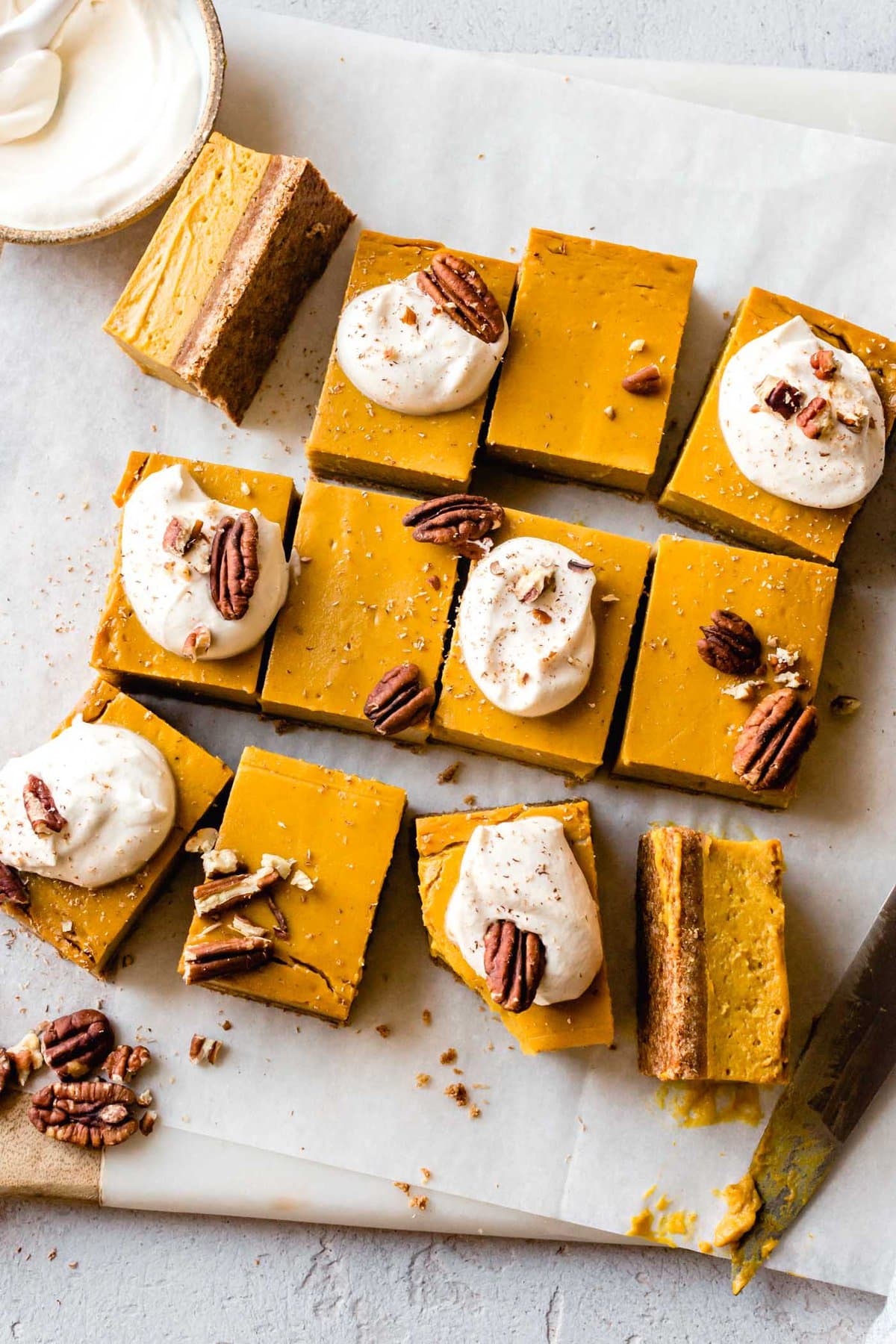 Thick pumpkin pie bars are arranged on a marble board and topped with dollops of cream and pecans