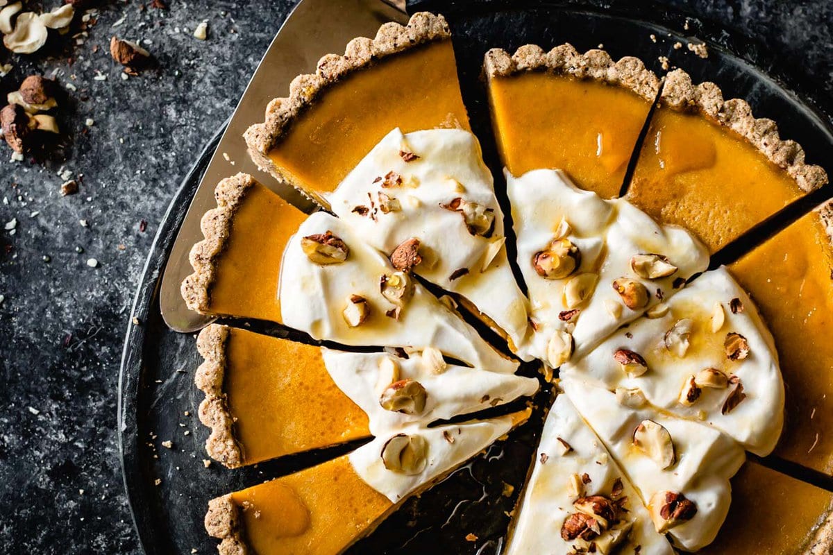 a close-up shot of a gluten-free pumpkin tart cut into wedges and topped with billows of cream, maple syrup, and chopped hazelnuts. 