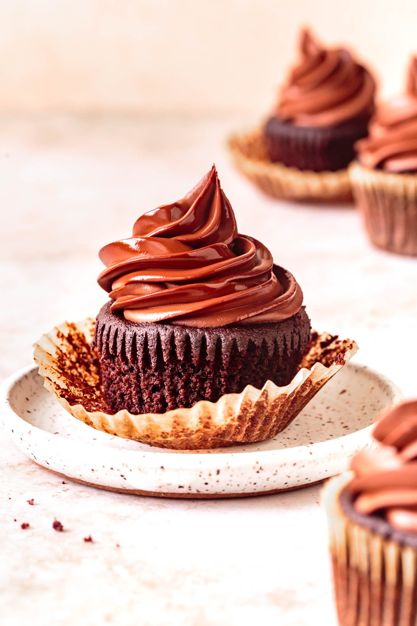 four chocolate cupcakes topped with glossy ganache are shown from the side on a warm speckled surface