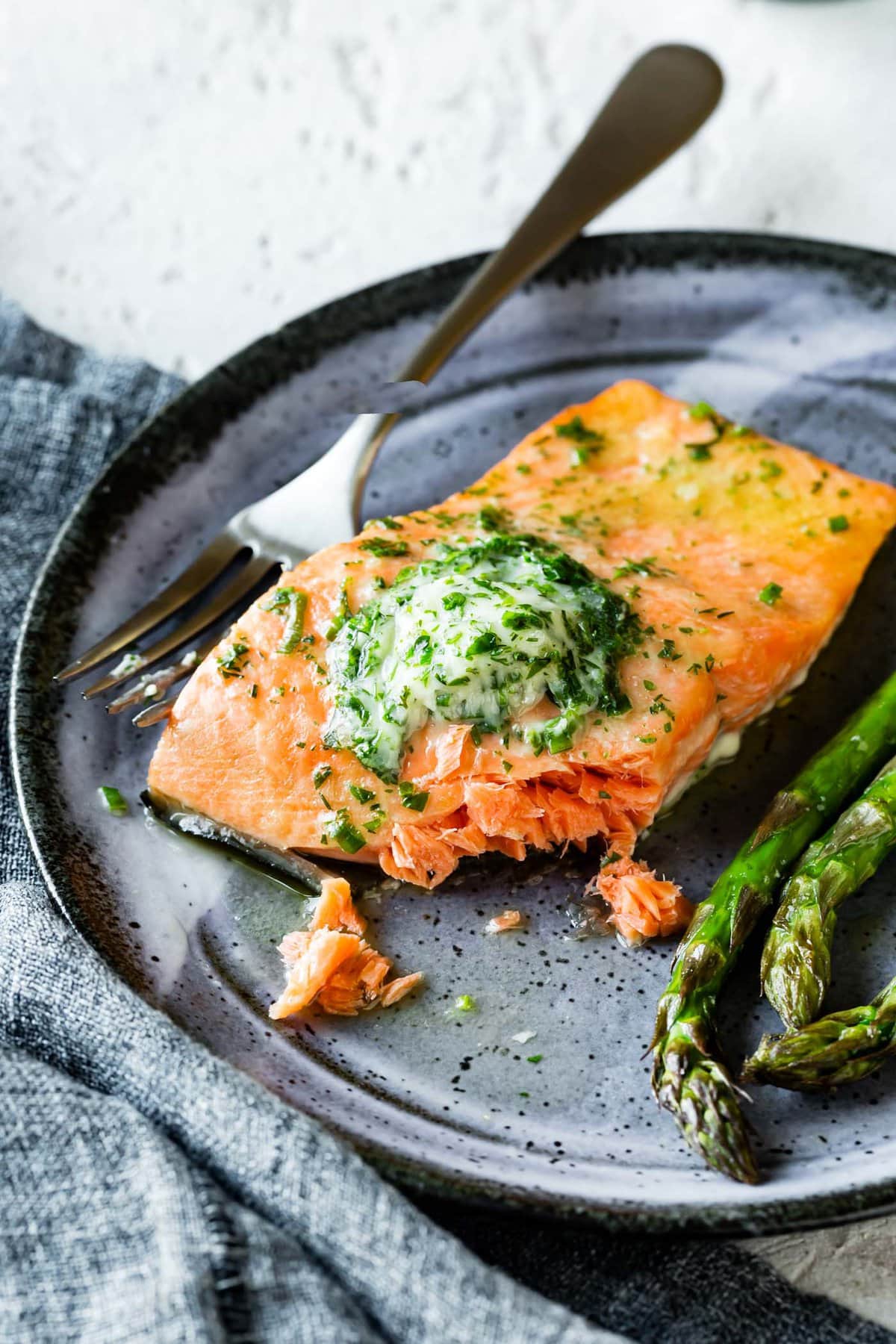 a piece of cooked salmon topped with herb butter and some asparagus are on a blue plate
