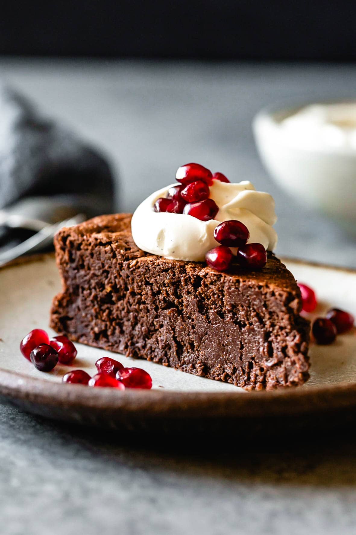 A slice of flourless chocolate hazelnut cake topped with pomegranate and cream, plated