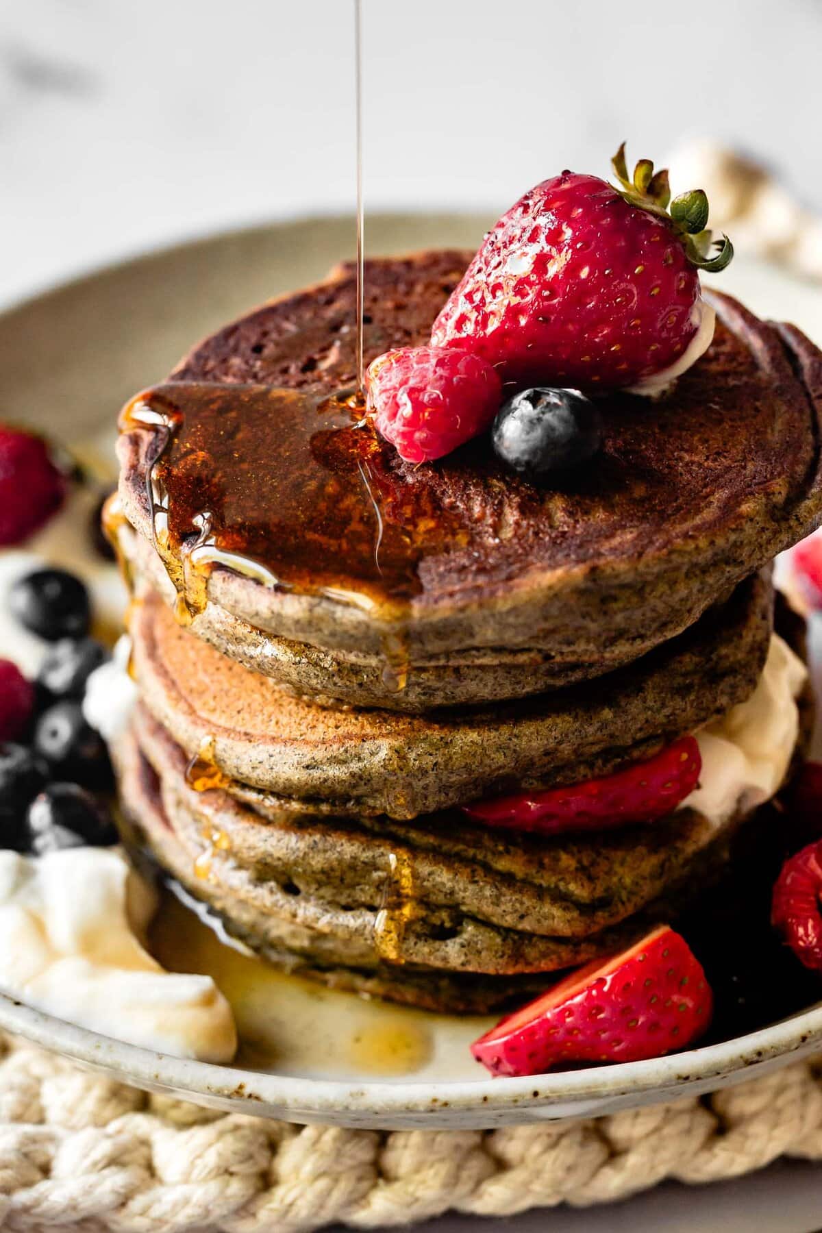 pouring syrup over a glorious stack of pancakes topped with berries and yogurt